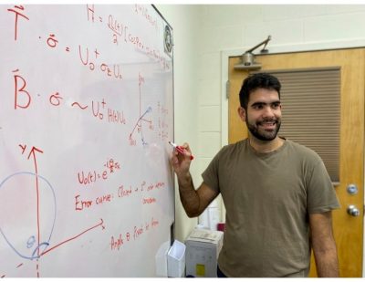 This phot shows a young man with black hair, beard, and eyebrows standing at a whiteboard writing equations with a red marker.