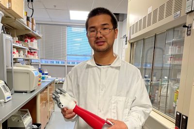 This photo shows a young Asian man with short black hair wearing a white lab coat and a pair of glasses. He is standing in a lab and holding a prosthetic forearm and hand.