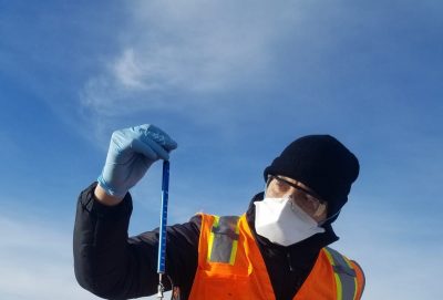 A photo of a man with a black beanie, safety glasses, a white safety mask, and a bright orange high visibility vest. He is holding a measuring device.