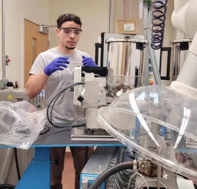 Photo of a young male PhD student in a lab with safety glasses and blue latex gloves waiting on something from a machine. 