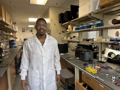 This photo shows a young Black man in a white lab coat standing in a laboratory and smiling at the camera. 