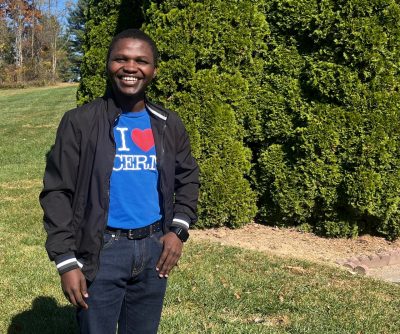 Gyang, a physics researcher, smiles for a picture on a sunny day with green trees and grass behind him.