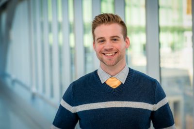 The image shows a smiling biostatistician posing for a picture in a hallway.