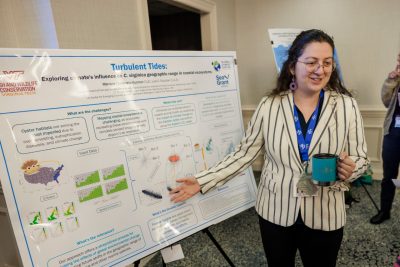 Mariana stands in front of her research poster, smiling and holding a cup of coffee as she talks about her research.