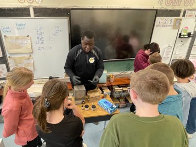 A graduate student stands at a table full of research supplies as a group of 8 elementary students listen in.