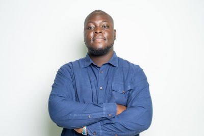 A man standing in front a blank, white wall, arms crossed, with a closed-mouth smile
