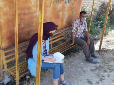 This photo shows a person clad in jeans, a blue shirt, a black head scarf, and a black mask sitting on a bench and taking notes on a clipboard. Also on the bench is a man in gray pants, a plaid shirt, and a gray or light blue face mask.