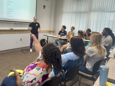 A female student standing in front of a projector screen displaying scientific words. She is smiling and gesturing with her hands as 10+ students sit in the classroom audience facing her. The professor is seated and emphatically pointing at the screen. 
