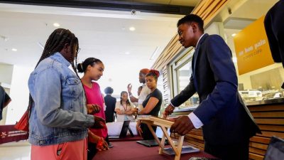 A young black male high school student in a suit stands behind a table smiling, pointing to a computer and bracing a wooden stand. Two young women stand opposite the table, focused on what he is presenting
