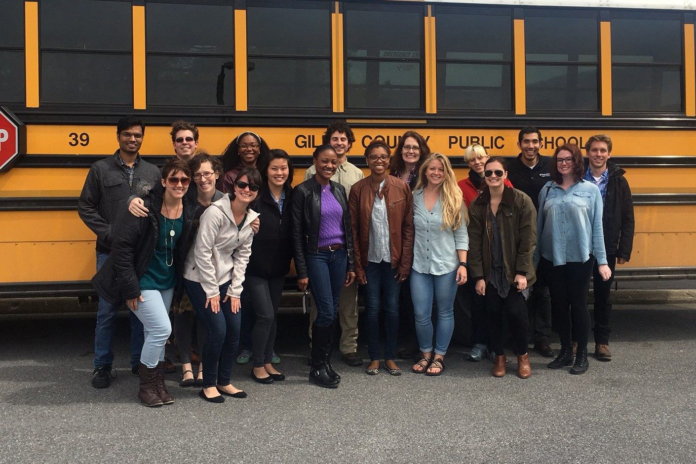 This photo shows 17 young adults (5 people of color) posed for a group photo in front of a yellow school bus with the words "Giles County Public Schools" on its side.