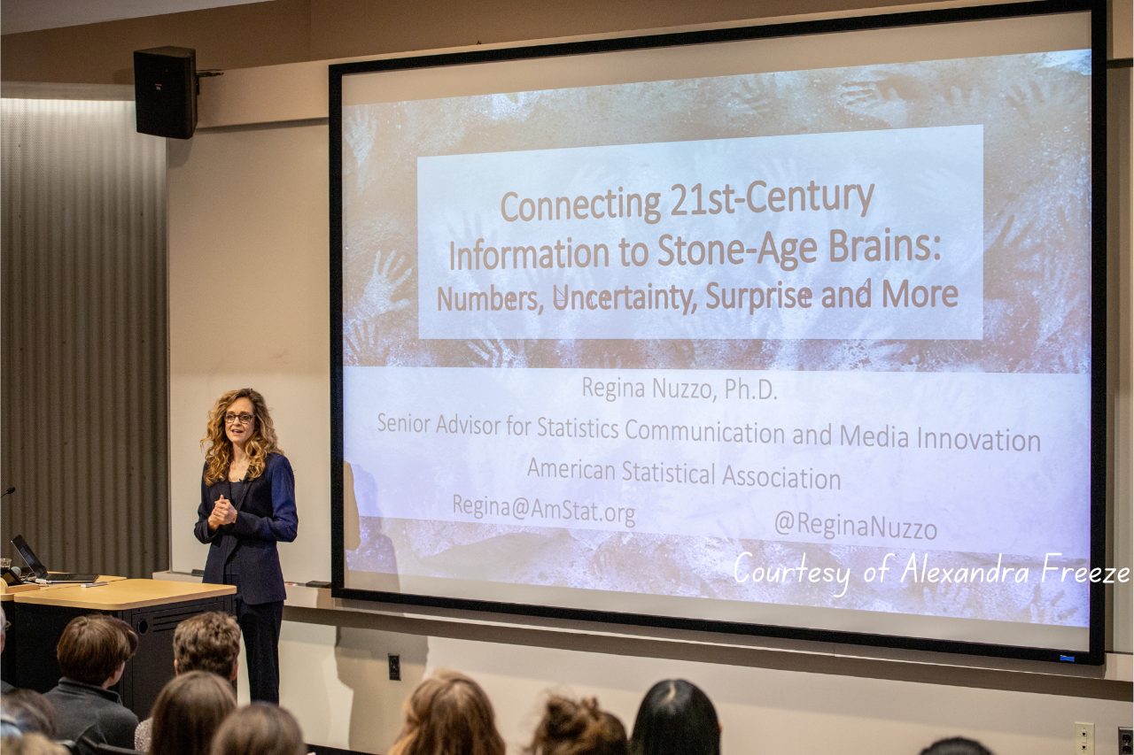 Presenter stands in front of large screen that displays presentation title on stone-age brains 