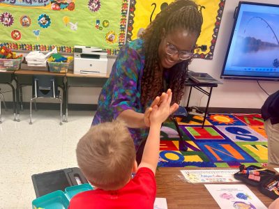 Woman smiles while high-fiving kindergarten boy sitting at desk with colorful rug and walls in the background.