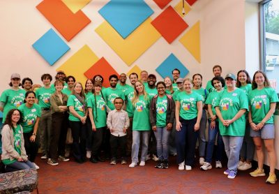 A large group of 25-30 people stand in rows in front of a wall decorated with colorful blocks. Most are wearing bright green t-shirts that read, "Flip the Fair" with colorful science lab beakers below "Flip the Fair"