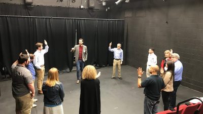 A group of people stand in a circle, led by Tobin (red shirt, grey blazer, jeans) at the back of the shot. A few people in the circle raise their hands. The curtain behind Tobin, the floors, and the walls are all black. 