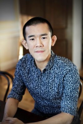 An Asian man with short hair looks straight at a camera while sitting in a chair. He is wearing a fun blue patterned shirt.