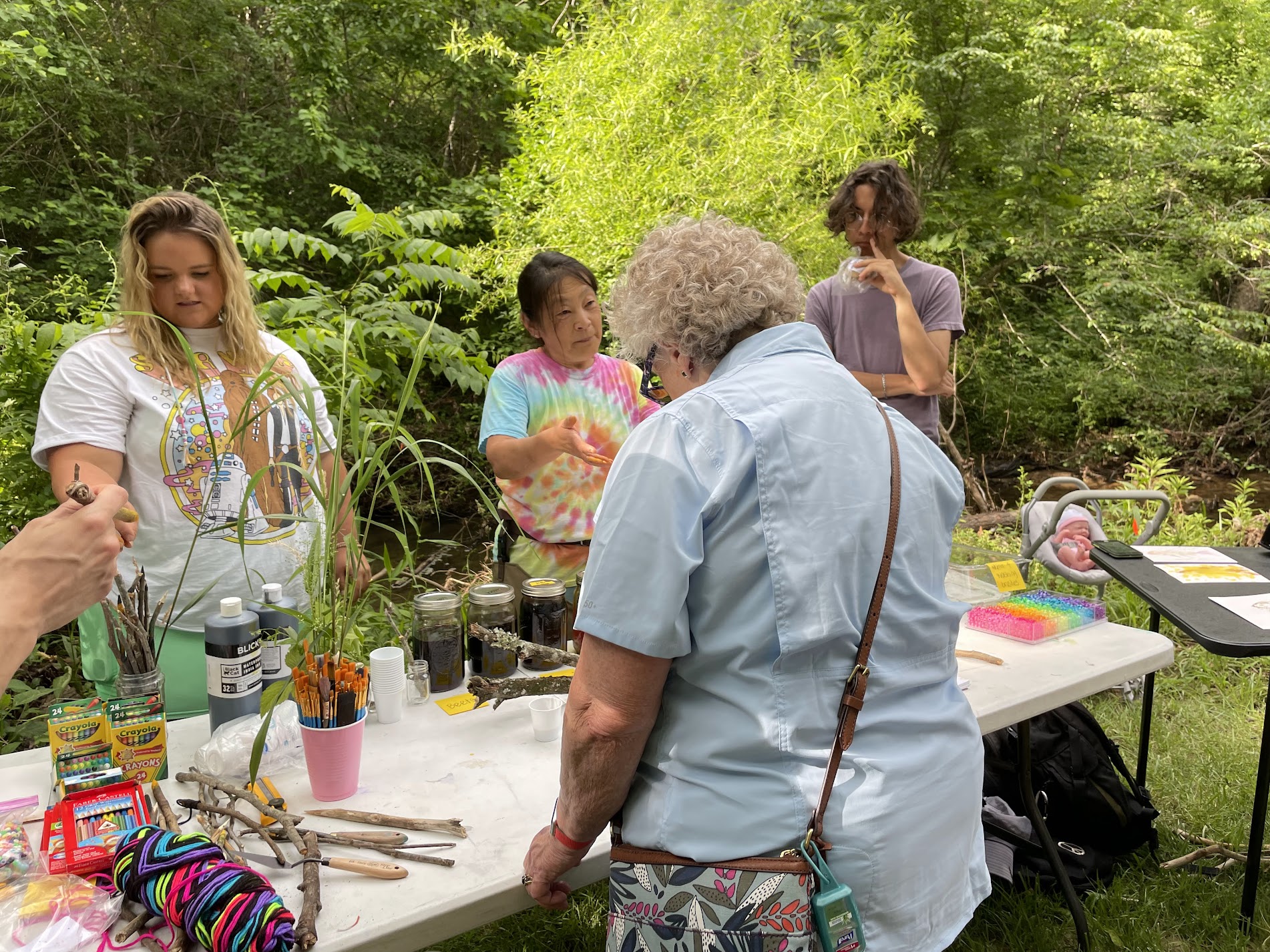 A group of diverse people stand around a white folding table. The table contains everything from paint paintbrushes, yarn, crayons, and sticks. In the center, an Asian woman in a tie-dye shirt speaks to an older woman in a short sleeve, light blue shirt. 