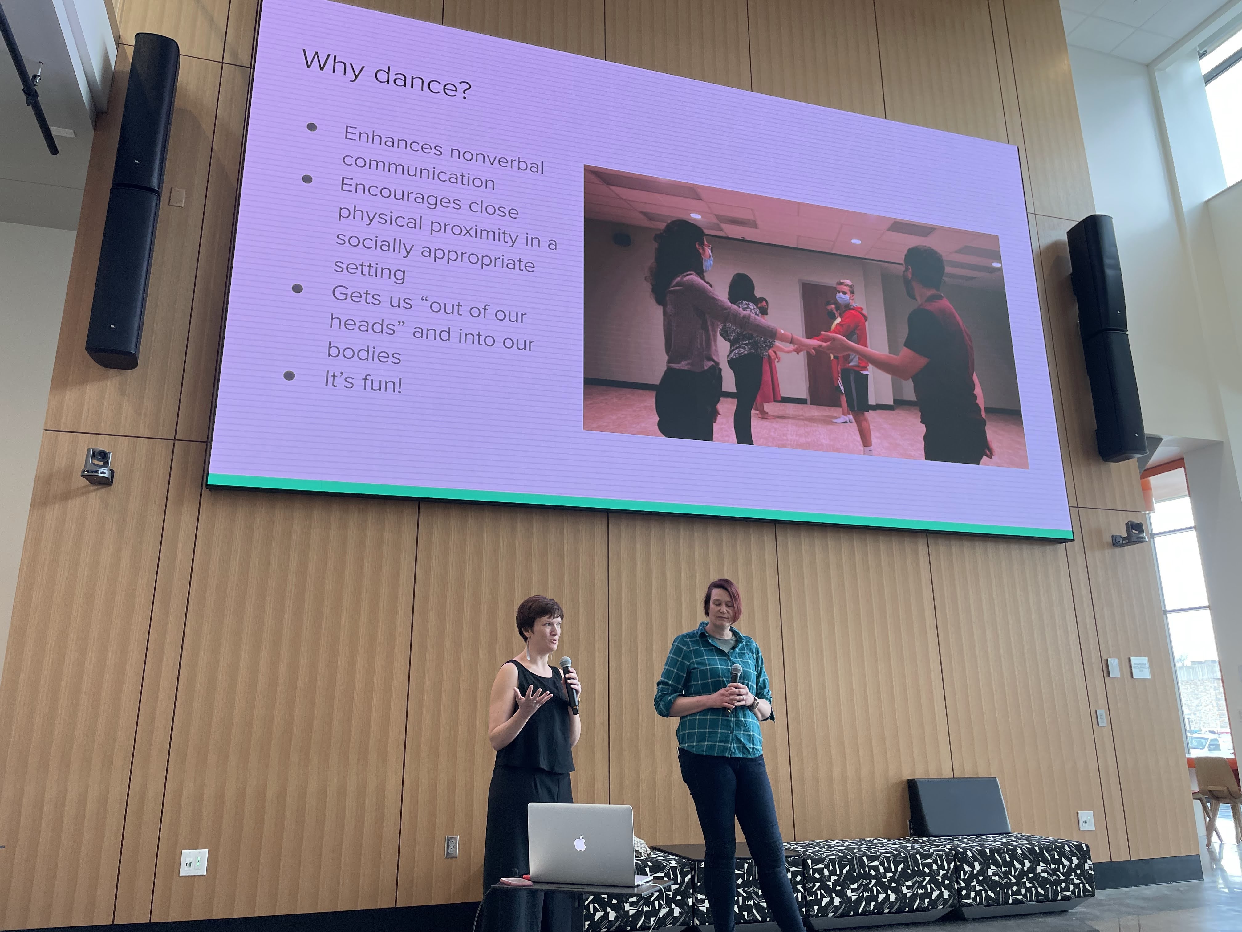 Two woman, one in a black dress and one in a blue shirt and jeans, stand under a presentation screen that reads ‘Why dance?’