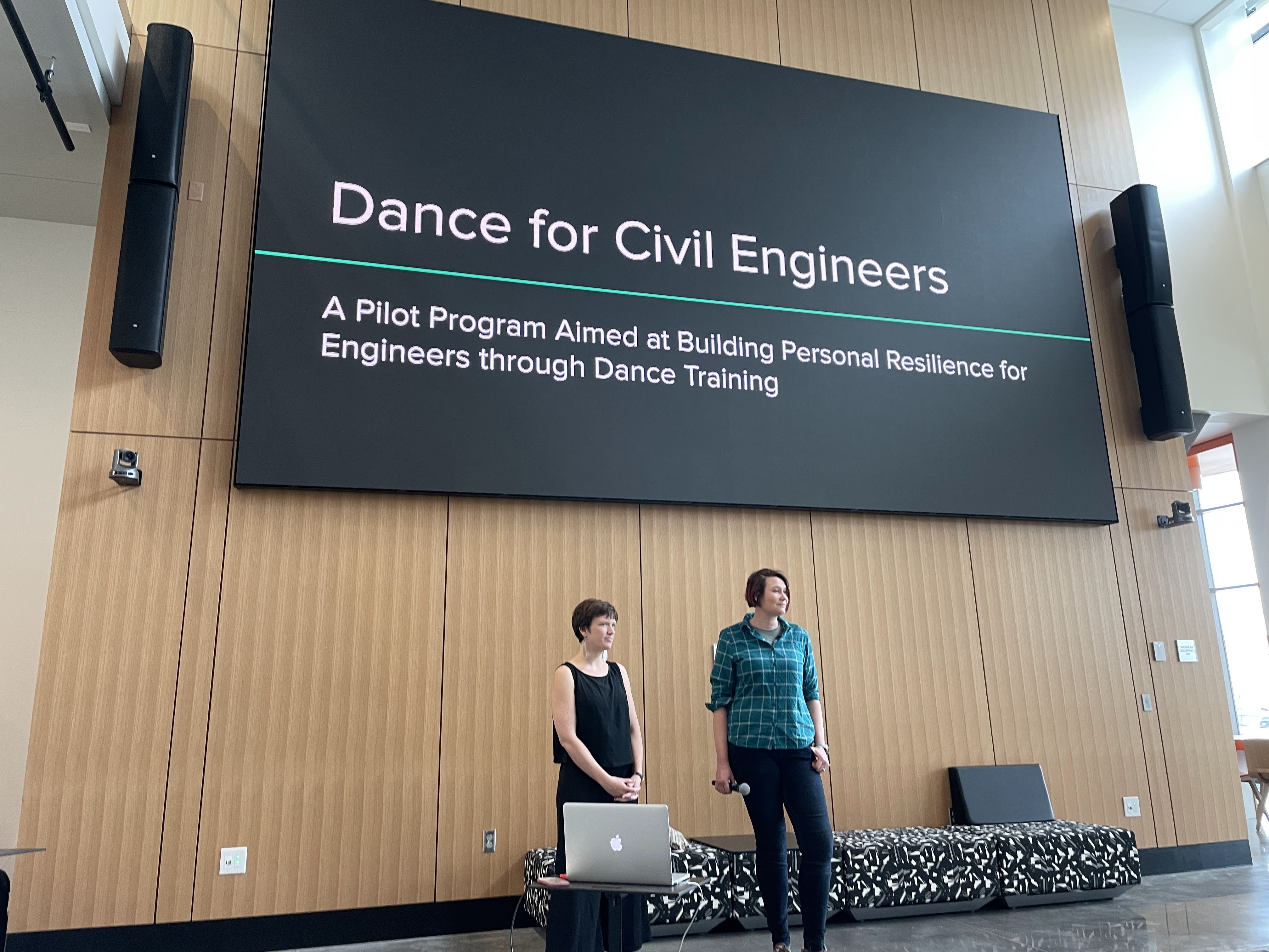 Two women, one wearing a black dress and the other wearing a blue shirt, stand under a presentation screen that reads “Dance for Civil Engineers.”