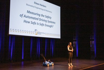 This photo shows a young white woman standing on a stage with a toddler's blue plastic push car behind her. Also behind her is a slide that reads "Eileen Herbers, Biomedial Engineering and Mechanics, Measuring the Safety of  Automated Driving Systems: How Safe is Safe Enough?"