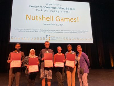 This photo shows five young people holding certificates and smiling at the camera with one older woman who does not have a certificate. Behind them is a large screen with the words Nutshell Games! on it.