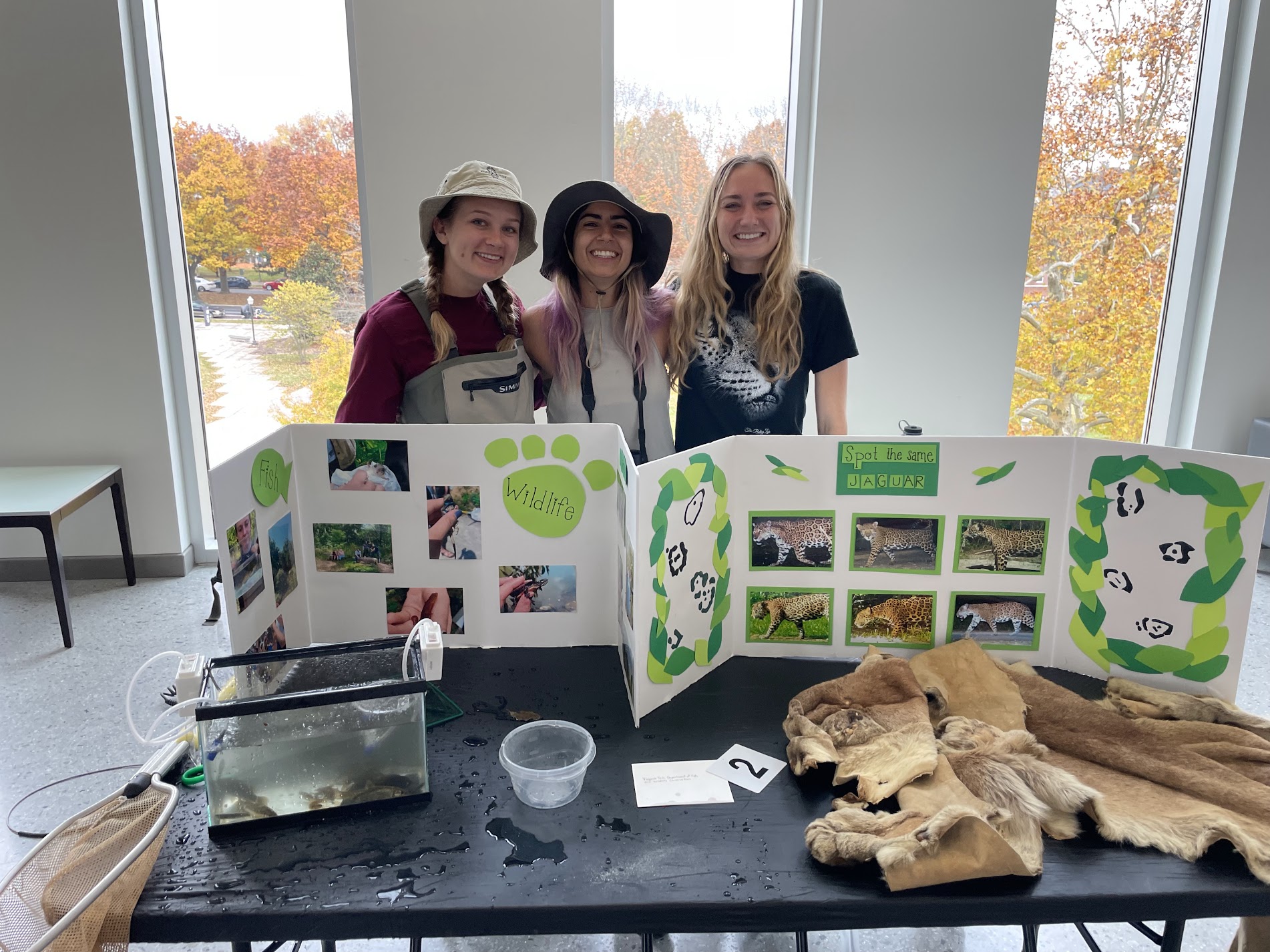 Three graduate students stand together and pose for a photo. On the table in front of them are two folding posters with photos of wildlife, an animal hide, and a small fish tank.