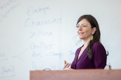 A White woman with medium-length brunette hair and glasses, wearing a dark purple blouse, stands at a podium in front of a white board with blue writing and story elements on it.