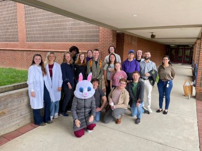 A group of students stand in the entryway of an elementary school. The two students on the far left are wearing lab coats, and in front of them, another student wearing a gray coat and red pants kneels down and wears a rabbit costume head.