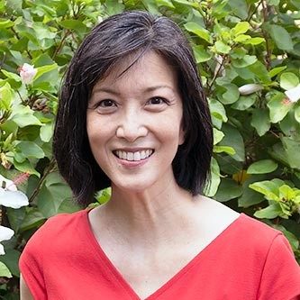 A middle-aged Asian woman with dark hair, wearing a red shirt, and leaves in the background smiles for a professional headshot.