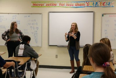 This photo shows two young white women standing in front of a classroom. The backs of a few middle school students' heads are visible. The wall is covered with whiteboards. 