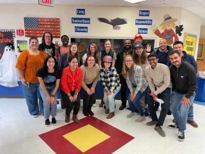 This photo shows 19 young adults of various skin colors and genders. Two are wearing hard hats. All are posed for a group photo with public school posters visible in the background.