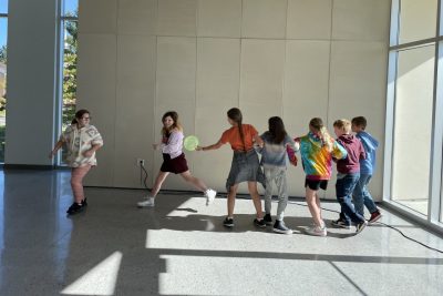 This photo shows 7 fifth graders running in an indoor space. Five have their arms linked and two are running free. 