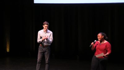 This photo shows a young white male in a light blue shirt and gray trousers and a young Black woman in a red top and black pants. She is holding a microphone and smiling and he is applauding. 