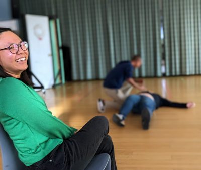 A participant looks into the camera, smiling, while two participants in the back mime CPR on each other.