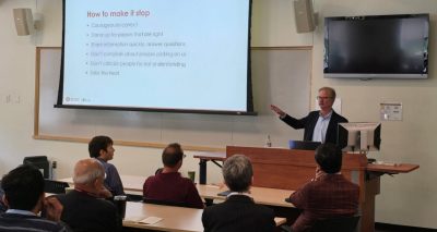 This photo shows a middle aged white man speaking from behind a lectern and gesturing toward the audience -- the backs of six people are visible in the foreground of the photo. On a screen behind him is projected a slide that says "How to make it stop."