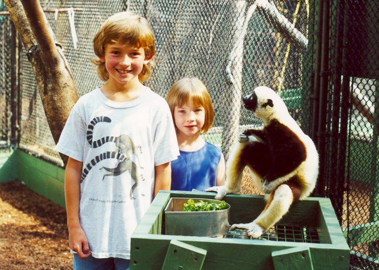 This photo shows two Caucasian light-haired children, probably about 7 and 4 years old, smiling at the camera. To their right is a strikingly patterned brown-and-white primate slightly smaller than the younger child. It is sitting on a green feeding tray containing a bowl of lettuce or some sort of greens. The animal is staring sintently at the older of the two children. All three seem to be inside a chainlink fence enclosure.