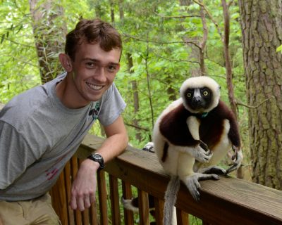 A young Causasian man poses and smiles at the camera, leaning on a deck rail. Sitting on the deck rail is a striking brown-and-white lemur with a black face, a coquerel's sifaka. In the immediate background is forest. 
