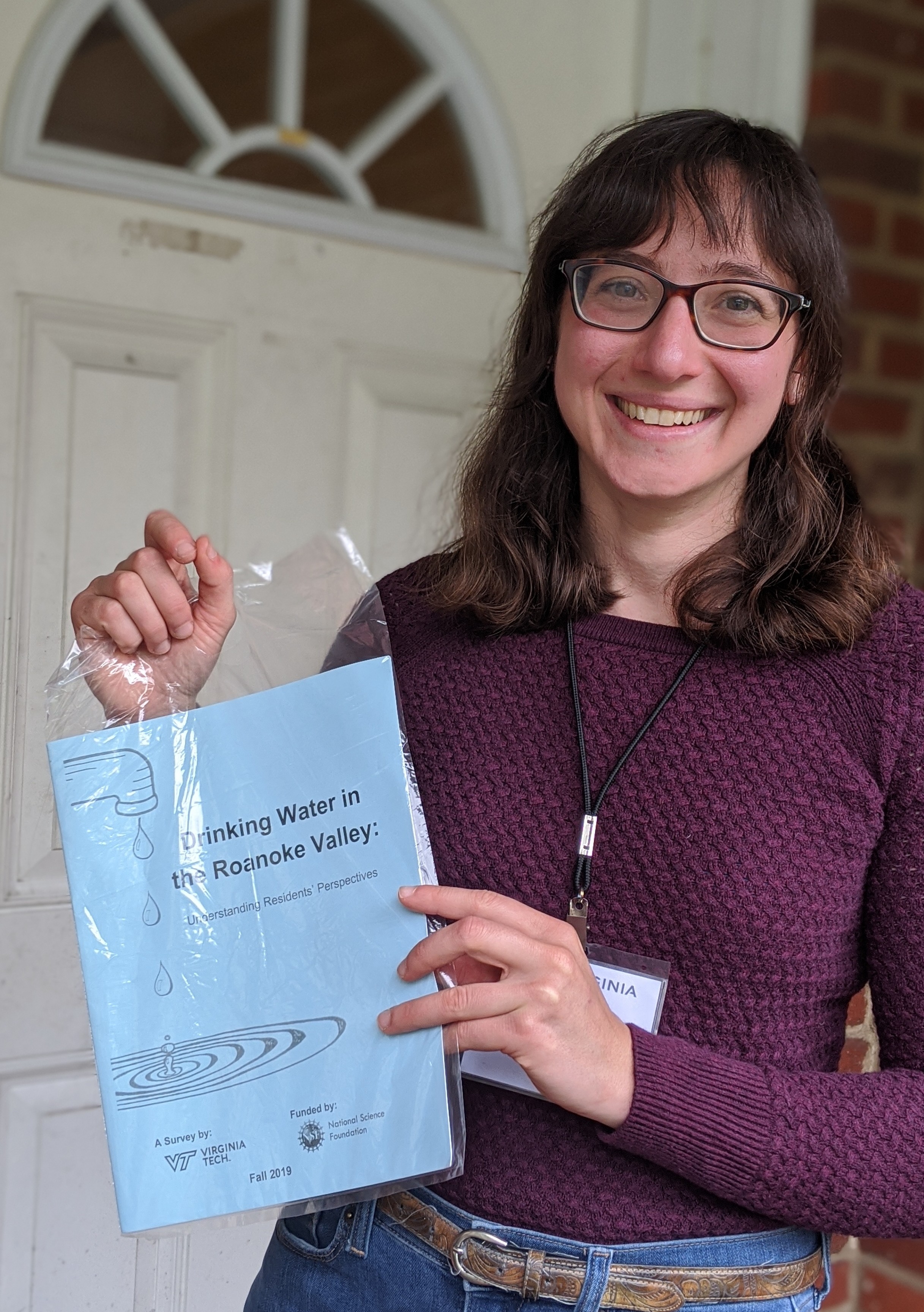 This photo shows a young Caucasian woman with shoulder-length brown hair, glasses, and a big smile. She is standing at the front door of a brick home holding a plastic bag with a blue book inside it titled "Drinking Water in the Roanoke Valley."