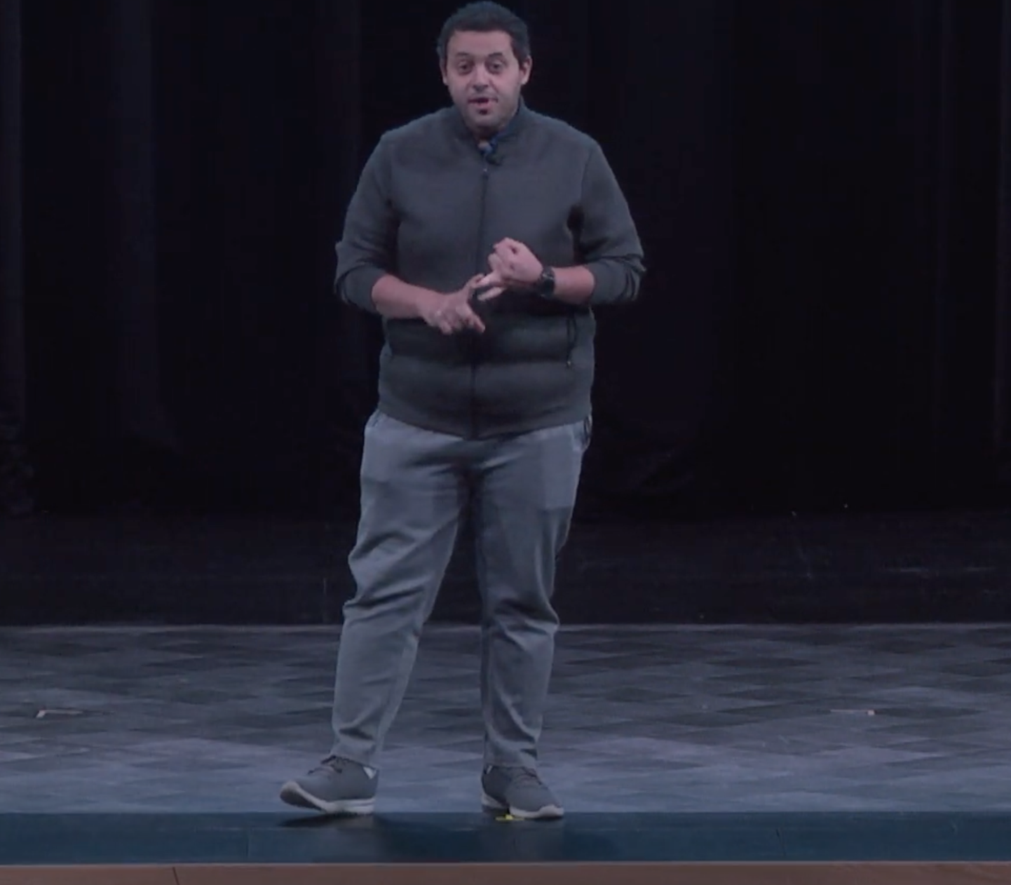 This photo shows the full body of a man on a stage speaking animatedly. He is wearing blue-gray pants, a gray sweater, and gray shoes. He has medium skin tone and dark short hair. 