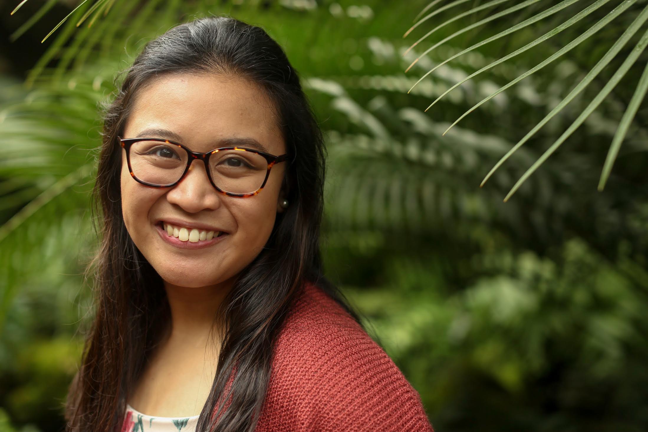 A head-and-shoulders shot of a young woman with medium-tone skin, long dark straight hair, glasses, and a big smile. The background is live plants, perhaps palm fronds.