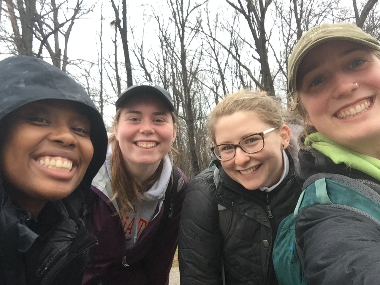 This photo shows the faces and upper torsos of four young women, one African American and three Caucasian, dressed for cold weather and smiling at the camera. In the background are leafless trees.