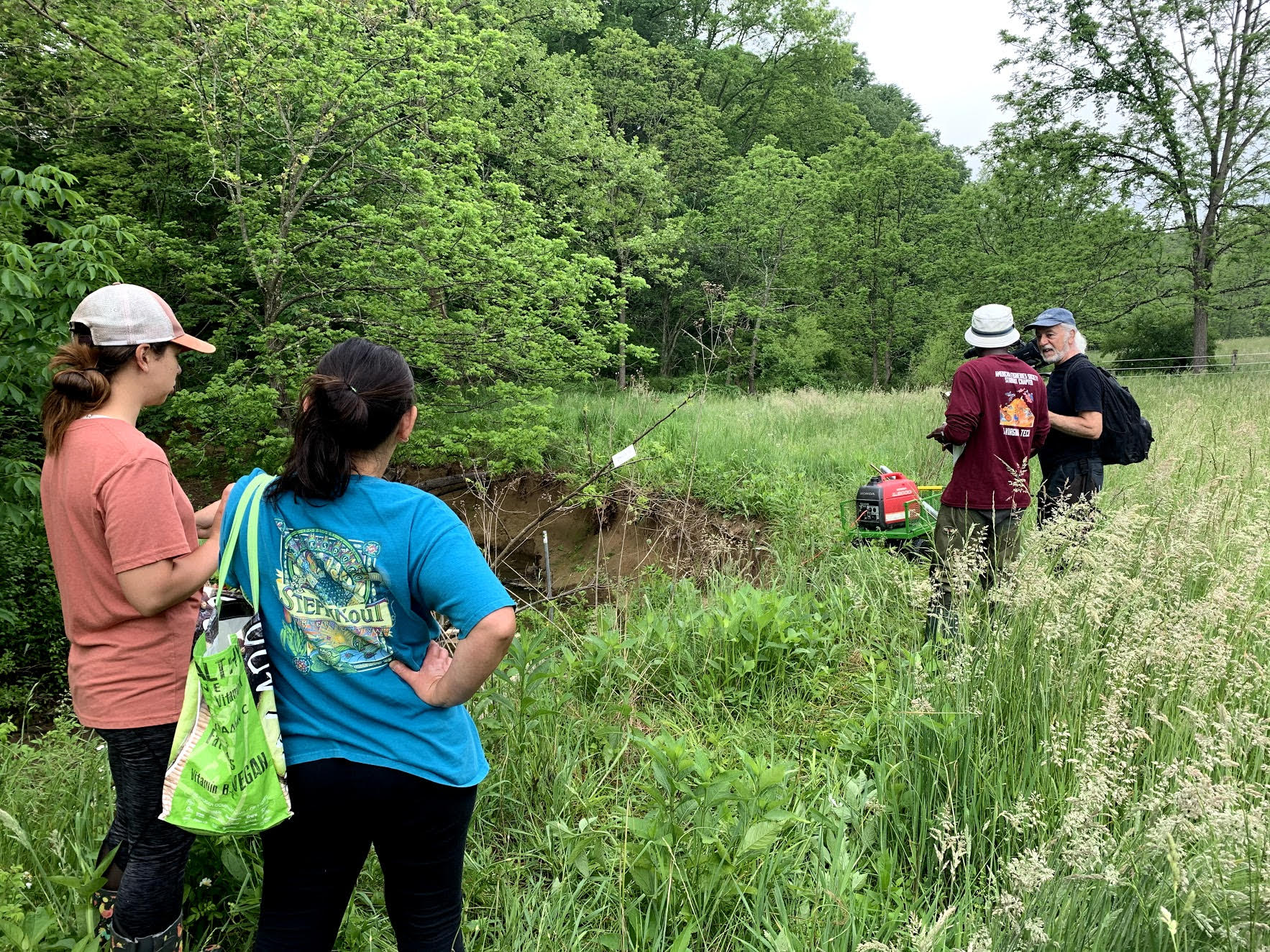 This photo shows in the foreground, and from the back, two women with hair in ponytails and farther from the camera two men, one from the back and one from the side, engaged in conversation. All are standing in a meadow near a creek bank. It appears to be high summer--everything is green and lush--and t-shirt colors include blue, salmon, maroon, and black.