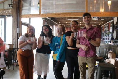 Five young adults (three white women, one Black woman, and one man) stand side by side smiling at the camera and holding up clear glass beakers with handles.