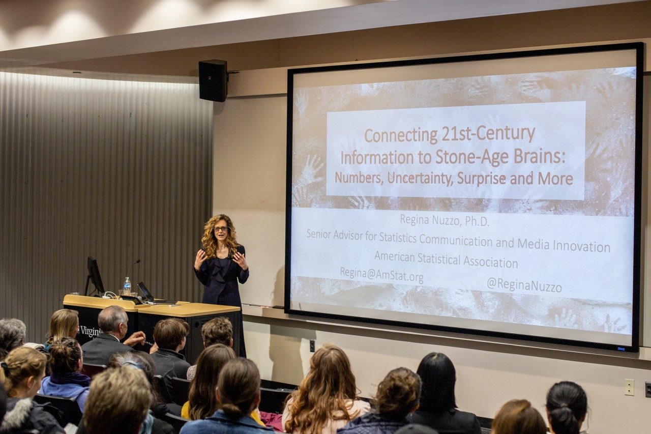 This photo shows an auditorium of people listening to a speaker. The speaker is a woman dressed in black with long curly brown hair. Behind her is a powerpoint slide with the title "Connecting 21st  Century Information to Stone-Age Brains: Numbers, Uncertainty, Surprise and More."