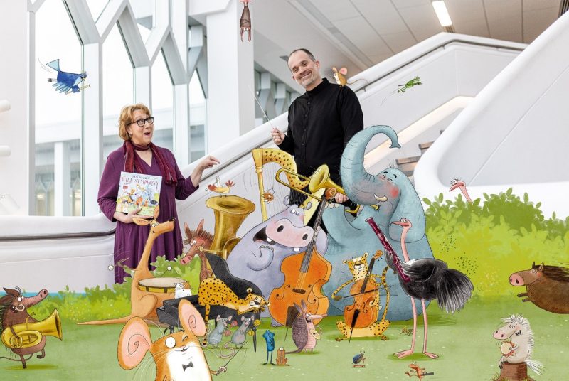 Patricia Raun , in a purple dress holding a book, and Derek Shapiro, dressed in black, stand behind a group of cartoon animals that are playing musical instruments.